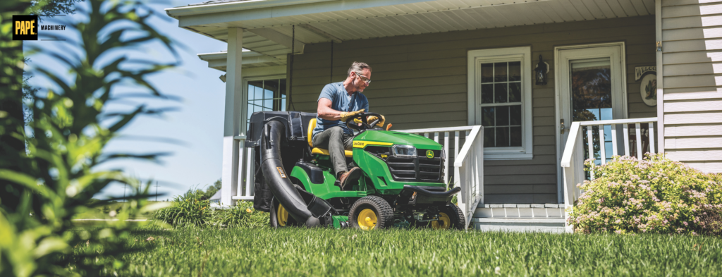 Man operating a green and yellow riding lawn mower on residential lawn in front of house with porch.