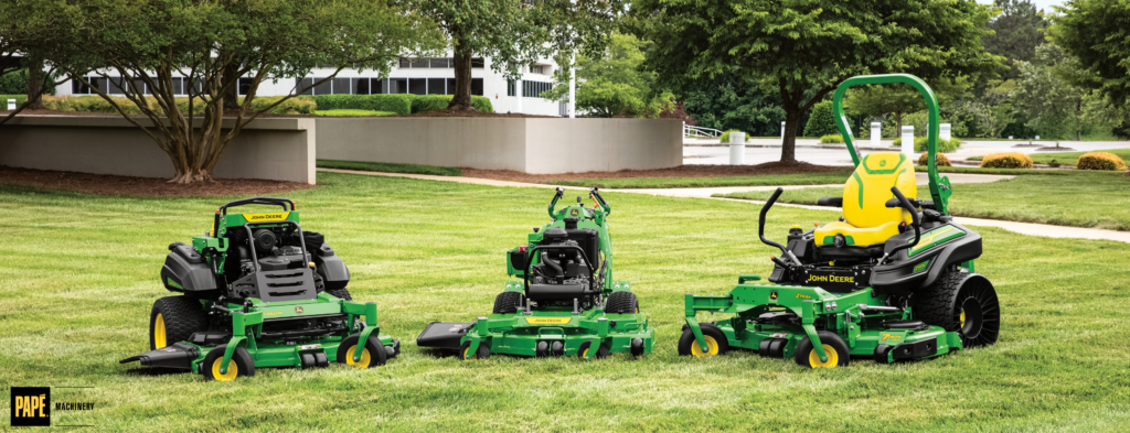 Three green and yellow lawn mowers displayed side-by-side on grass with buildings and trees in the background.