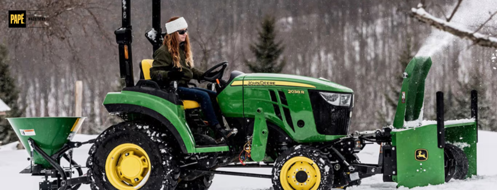 Person operating green and yellow tractor with snow removal equipment attached in winter landscape.