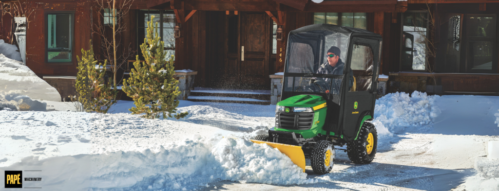 Operator in green and yellow snow removal vehicle with plow blade clearing snow from residential driveway.