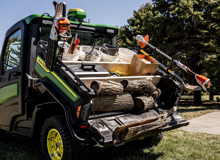 Green and black pickup truck with open bed displaying firewood logs, chainsaws, and forestry equipment organized for transport.