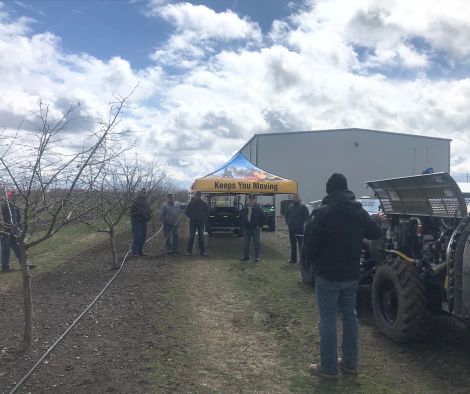 Outdoor farm equipment event with attendees gathered around machinery and a tent with yellow awning in an orchard setting.