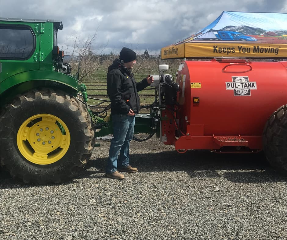 Man operating fuel transfer equipment between a green tractor and red tank trailer on a gravel lot.