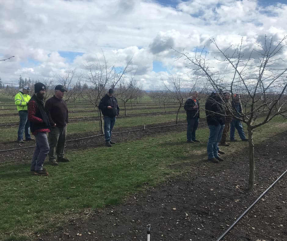 A group of people standing in a bare orchard during a daytime training or tour, with leafless trees and mountains visible in the background.