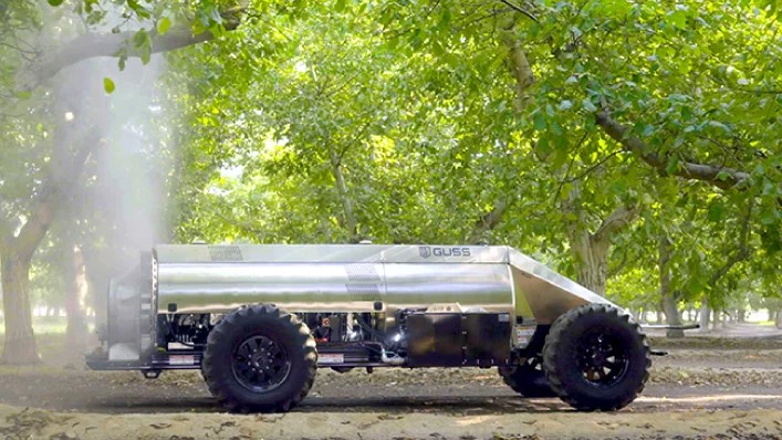 Polished metal tanker trailer with large black wheels parked under tree canopy on dirt ground.