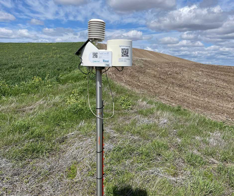 Weather monitoring station mounted on a pole in an agricultural field with green crops on the left and tilled soil on the right.