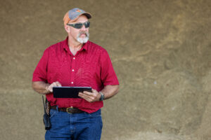 Man in red shirt holding tablet device while standing outdoors against tan wall.