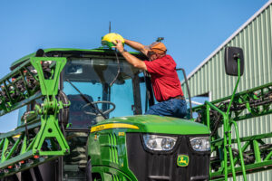 Operator in red shirt and hard hat placing yellow helmet on top of green agricultural sprayer cab in field.