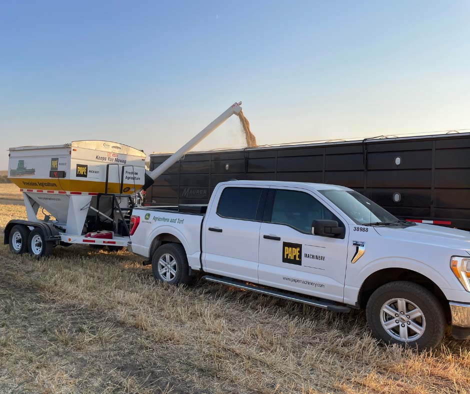 White pickup truck towing a grain cart auger with extended boom toward a dark agricultural trailer in a harvested field.
