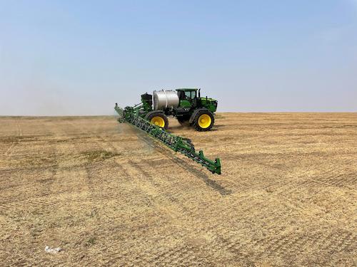 Green tractor with spray boom applicator spraying herbicide across a plowed field.