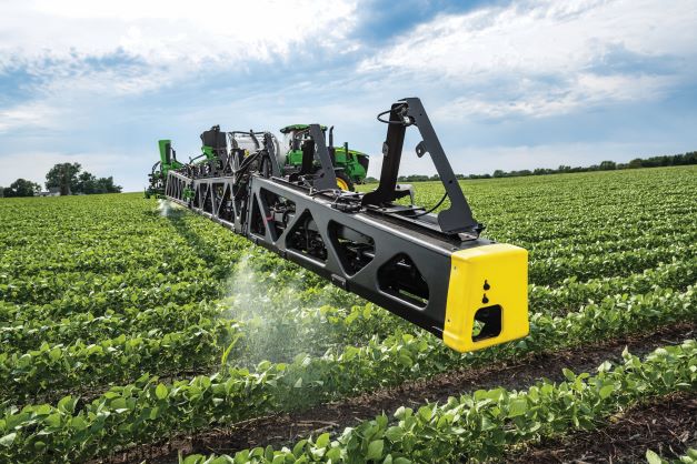 Green tractor with spray equipment applying liquid pesticide or fertilizer across a soybean field.