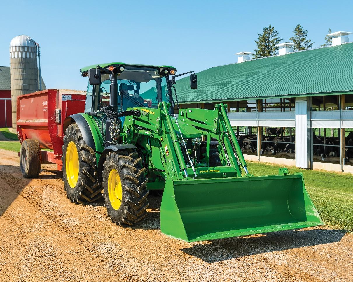 Green farm tractor with front loader and red wagon on rural farm property with barn and silo.
