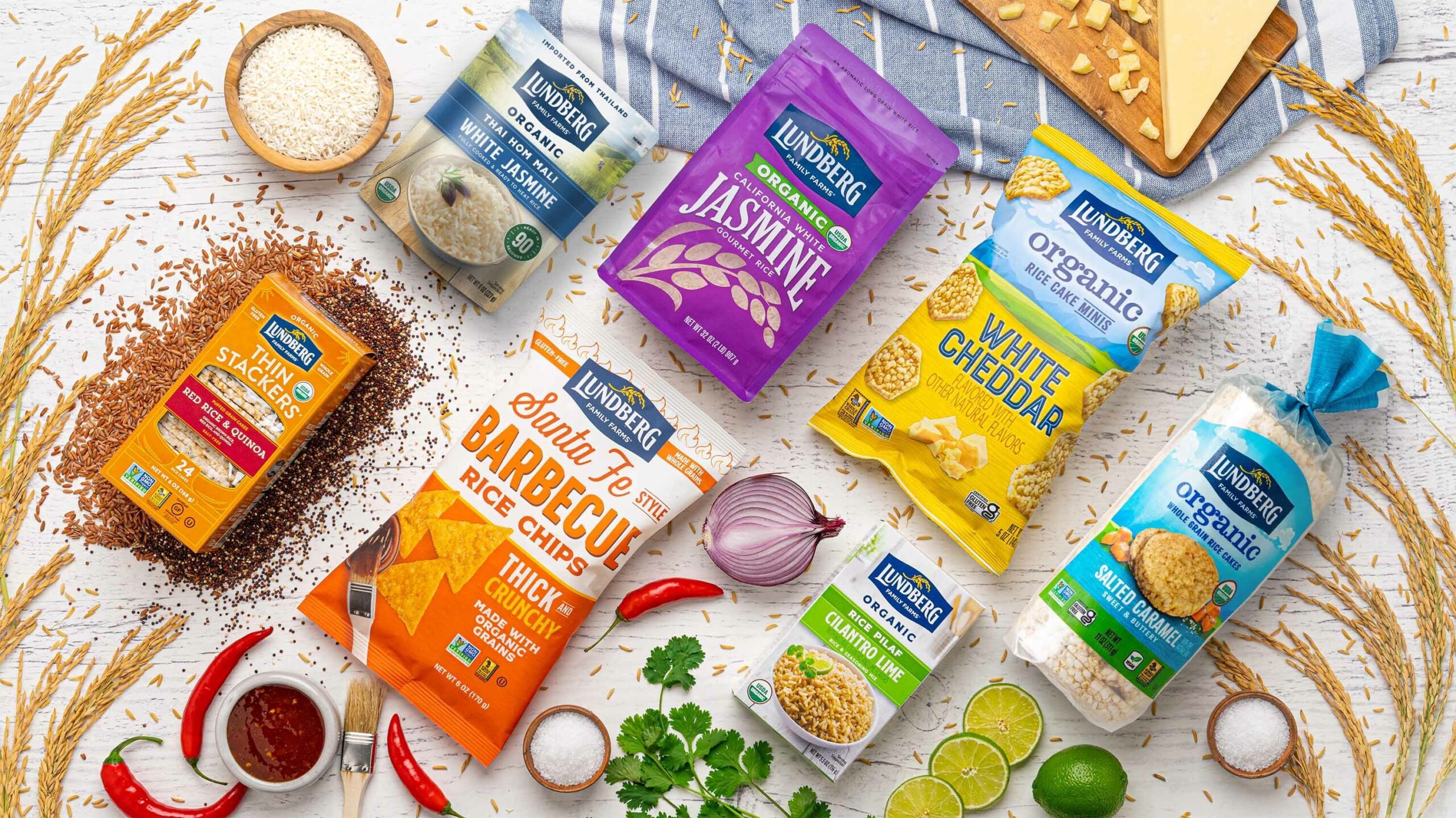 Lundberg organic rice and grain products arranged with fresh ingredients including red chili peppers, lime, cilantro, onion, and cheese on a white wooden surface.