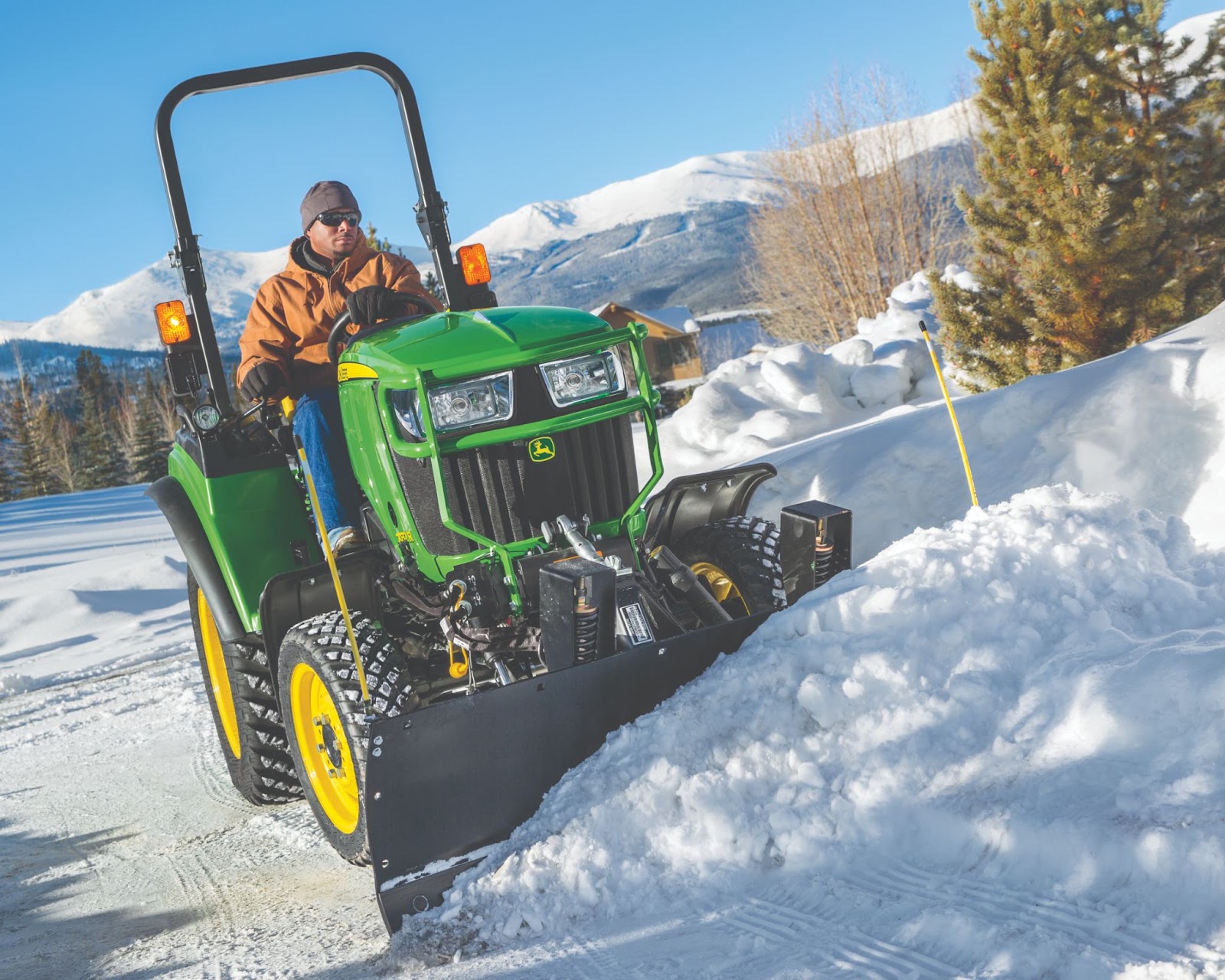 This image features a man operating a John Deere 2032R Compact Tractor equipped with a front blade attachment, clearing snow from a driveway or rural road in a winter landscape. The operator is wearing a brown winter jacket, gloves, sunglasses, and a beanie, suitable for cold weather conditions. The tractor is fitted with turf-friendly, deep-tread tires, likely for enhanced traction on snow and ice. The background showcases a snow-covered mountainous region with pine trees and a clear blue sky, indicating a cold but sunny winter day. The bright yellow snow stakes on the side of the road help mark boundaries for snow removal. The image conveys winter utility and efficiency, demonstrating the versatility of John Deere compact tractors for seasonal maintenance and snow management in residential, commercial, or rural settings.