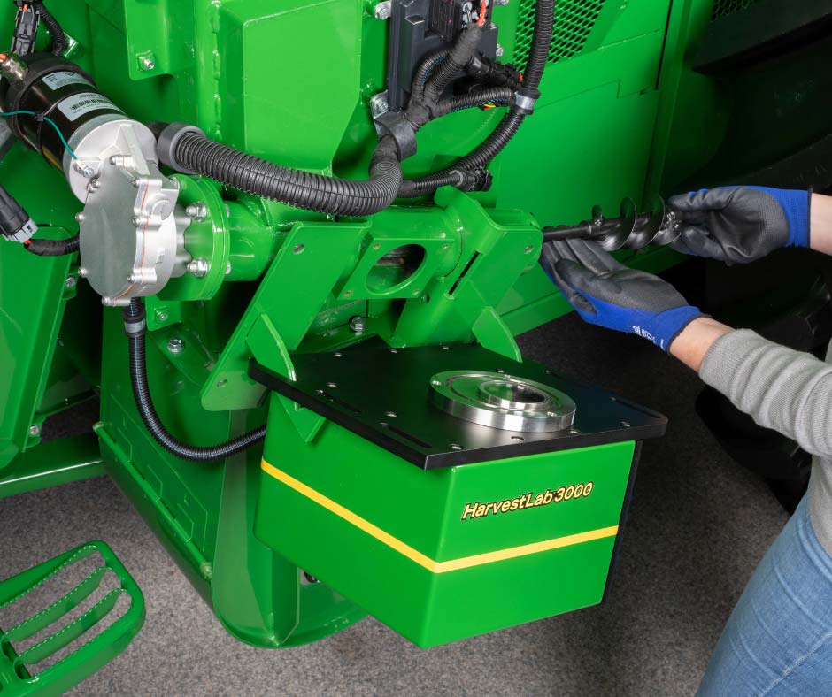 Technician in work gloves installing or servicing a stainless steel component on a green agricultural machine with hydraulic hoses and electrical connections visible.